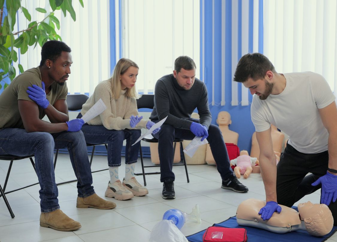 Group of students participating in a CPR class, watching an instructor perform chest compressions on a mannequin.